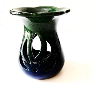 May include: A green and blue ceramic oil burner with a decorative cutout design. The burner has a wide, flat top and a round base.