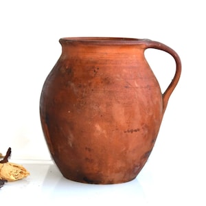 Op de afbeelding: Een terracotta kan met een rond lichaam en een enkele handgreep. De kan heeft een rustieke, verweerde uitstraling.