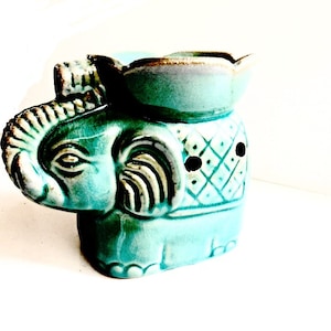 May include: A teal blue ceramic elephant oil burner with a decorative design. The burner has a small bowl on top for wax melts or essential oils.