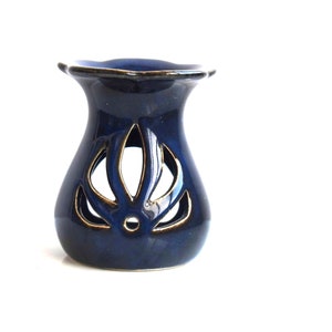 May include: A dark blue ceramic essential oil diffuser with a decorative floral cutout design. The diffuser has a flared shape with a scalloped top and gold trim. The product is designed to hold essential oils and release their fragrance.