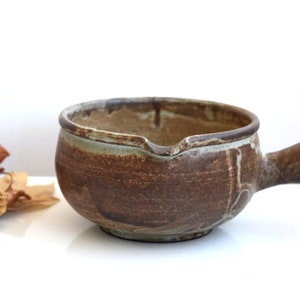 May include: A brown ceramic serving dish with a handle. The dish has a light grey glaze and a rustic, hand-thrown look.
