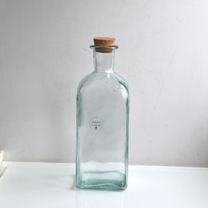 Könnte beinhalten: Eine klare Glasflasche mit einem Korkverschluss. Die Flasche hat eine rechteckige Form und ein Etikett mit der Aufschrift "Vidrio" und einem kleinen "M" darunter.