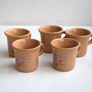 Puede incluir: Cinco tazas de cerámica color beige con asas, cada una adornada con un delicado diseño floral en tonos rosa y morado. Las tazas tienen una forma ligeramente acampanada y están sobre una superficie blanca.