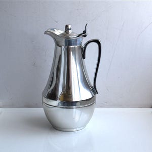Può includere: Una caraffa in metallo color argento lucido con corpo affusolato, base ampia e arrotondata e manico curvo. La caraffa presenta un coperchio incernierato con una piccola leva per l'apertura, un beccuccio pronunciato e una finitura metallica liscia.