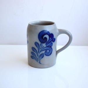 Op de afbeelding: Een grijze keramische bierkan met een blauw bloemenmotief. De kan heeft een handvat en een ronde basis.