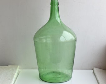 Puede incluir: Una gran botella de vidrio verde transparente con una boca ancha y un fondo redondeado. La botella está vacía y tiene un aspecto vintage.
