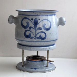 Könnte beinhalten: Ein blauer und weißer Keramik-Fonduetopf mit Blumenmuster. Der Topf steht auf einem Metallständer mit einem kleinen Kupferbrenner.