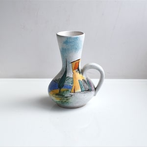 Puede incluir: Un jarrón de cerámica blanca con asa, que presenta una escena pintada a mano de una casa, árboles y un lago azul. El jarrón tiene un estilo rústico y vintage.