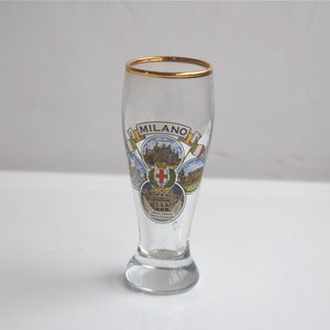 May include: A clear glass shot glass with a gold rim and a printed design featuring the word "MILANO" and images of buildings.