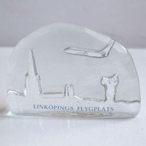 May include: A clear glass paperweight with a raised design of a church and an airplane. The text "LINKÖPINGS FLYGPLATS" is etched on the bottom.