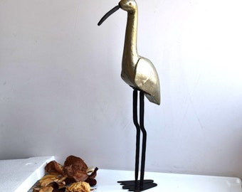 Op de afbeelding: Een gouden houten vogelsculptuur met zwarte metalen poten. De vogel heeft een lange nek en een lange snavel.