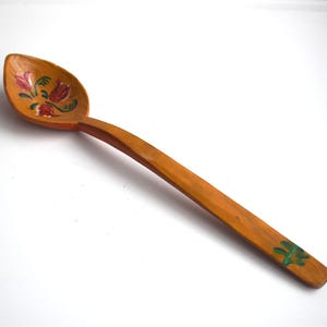May include: A wooden spoon with a hand-painted floral design in shades of red, green, and yellow. The spoon has a long, slender handle and a rounded bowl.