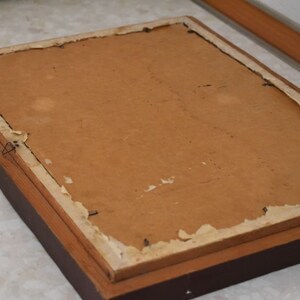 May include: The reverse of a framed picture, featuring a brown wooden frame. The backing is a light brown cardboard, with paper peeling away at the edges. A wire is attached for hanging.