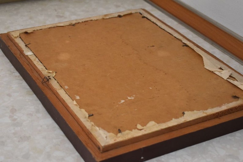 May include: The reverse of a framed picture, featuring a brown wooden frame. The backing is a light brown cardboard, with paper peeling away at the edges. A wire is attached for hanging.