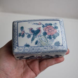 Könnte beinhalten: Eine kleine, weiße Porzellanbox mit einem blauen und grünen Blumendesign. Die Box zeigt zwei Vögel, die auf einem Zweig mit rosa Blüten sitzen.