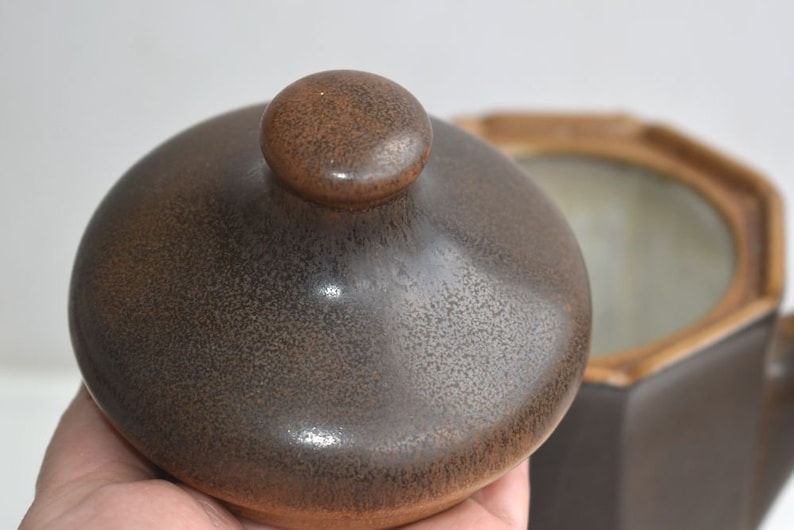 Może przedstawiać: Brązowa ceramiczna pokrywka z fakturowaną powierzchnią i zaokrąglonym uchwytem na g&oacute;rze. Pokrywka jest do pojemnika do przechowywania.