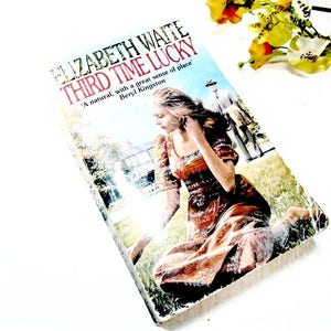 May include: A paperback book titled "Third Time Lucky" by Elizabeth Waite. The cover features a woman in a brown dress sitting in a field with a man in a suit walking away in the background.