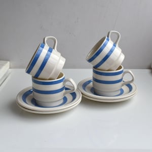 Puede incluir: Dos juegos de tazas y platillos de cerámica apilados, cada uno con una base blanca y rayas azules horizontales. Las tazas tienen un diseño escalonado cerca de la base y un asa curva, mientras que los platillos a juego muestran un patrón de rayas azules.