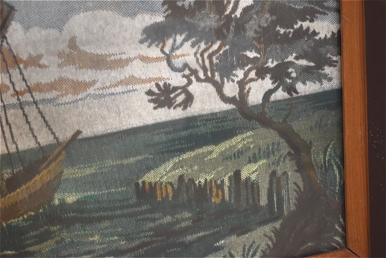 May include: A framed tapestry showing a coastal scene with a sailboat, a tree, and a shoreline. The artwork uses brown, green, and blue hues to create a tranquil landscape.