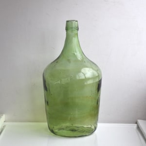Op de afbeelding: Een grote, groene glazen fles met een brede opening en een rond lichaam. De fles is leeg en heeft een licht getextureerd oppervlak.