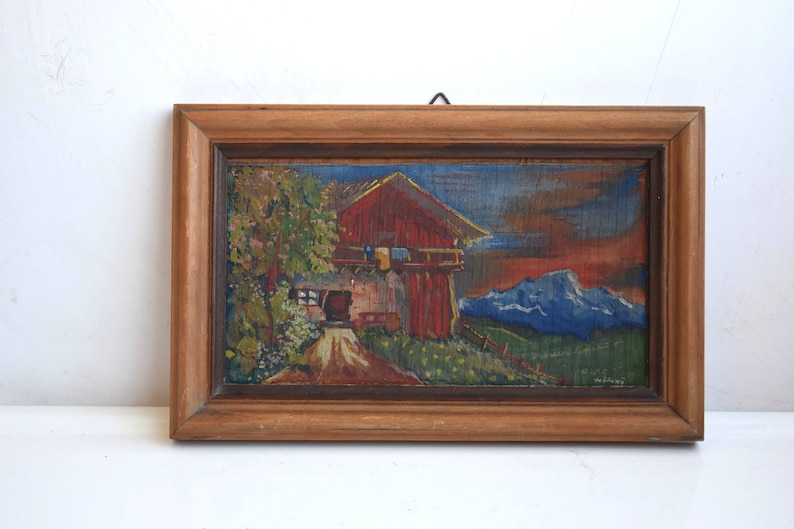 K&ouml;nnte beinhalten: Ein gerahmtes Gem&auml;lde, das ein rotes Haus mit blauem Dach und wei&szlig;en Verzierungen zeigt. Das Haus steht vor dem Hintergrund gr&uuml;ner H&uuml;gel und blauer Berge. Das Gem&auml;lde ist signiert mit "E.W.T. Melzer".
