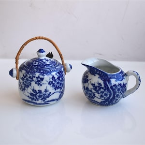 Può includere: Un servizio da tè in porcellana blu e bianca con una piccola teiera con coperchio e manico in vimini e una lattiera. Entrambi i pezzi sono decorati con un motivo floreale. Il servizio da tè è su una superficie bianca.