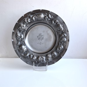 May include: A round grey metal plate featuring an embossed floral border with swirling leaf patterns. The centre of the plate displays an engraved monogram with the letters RC and the date 19 11 47 inscribed directly below the central design.