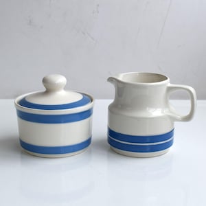 May include: A cream-colored ceramic sugar bowl with a lid and a matching creamer pitcher. Both pieces feature two horizontal blue stripes encircling the body. The sugar bowl has a rounded lid with a small knob handle, and the creamer has a spout.