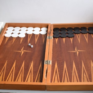 Puede incluir: Un tablero de backgammon de madera con un diseño de incrustaciones marrón y naranja. El tablero está abierto y muestra fichas blancas y negras. Dos dados blancos están en el tablero.