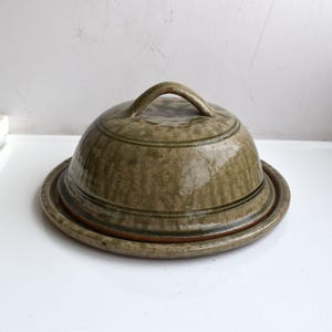 Peut inclure: Un beurrier rond en céramique vert olive avec un couvercle bombé doté d'une poignée incurvée. La surface présente un vernis texturé et moucheté avec de fines bandes horizontales entourant le dôme, reposant sur une base circulaire assortie.