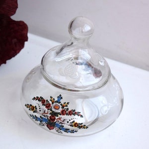 K&ouml;nnte beinhalten: Ein klares Glasgef&auml;&szlig; mit Deckel, verziert mit einem Blumendesign in Rot, Blau, Gelb und Gr&uuml;n. Das Gef&auml;&szlig; hat ein erhabenes, strukturiertes Design auf Deckel und K&ouml;rper.