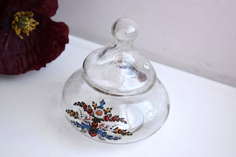 K&ouml;nnte beinhalten: Ein klares Glasgef&auml;&szlig; mit Deckel, verziert mit einem Blumendesign in Rot, Blau, Gelb und Gr&uuml;n. Das Gef&auml;&szlig; hat ein erhabenes, strukturiertes Design auf Deckel und K&ouml;rper.