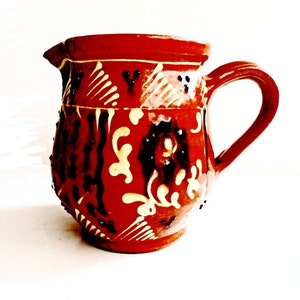 Op de afbeelding: Een rode kleipot met een wit en zwart bloemenontwerp. De pot heeft een handvat en een rond lichaam.