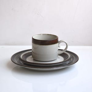 Pode incluir: Uma caneca de cerâmica com borda e alça marrons está sobre uma pilha de dois pratos combinando. A louça tem um esquema de cores branco e marrom. A louça está em um fundo branco.