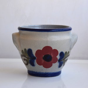 Op de afbeelding: Een witte keramische pot met blauwe en rode bloemenmotieven. De pot heeft twee handvatten aan de zijkanten.