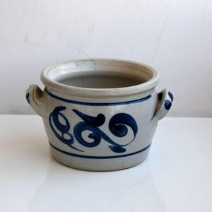 Op de afbeelding: Een lichtgrijze keramische pot met twee handvatten aan de zijkanten en een met de hand geschilderd donkerblauw wervelpatroon aan de buitenkant. De pot heeft een brede ronde opening en een dikke rand, met een horizontale blauwe lijn onderaan.