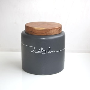 May include: A dark grey ceramic container with a wooden lid. The word "Zwiebeln" is written in white script across the container. The container is designed for kitchen storage.