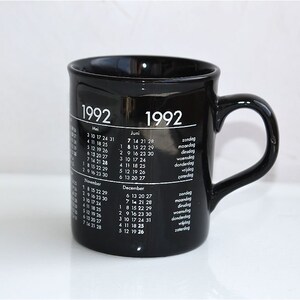 May include: Black ceramic mug with a white calendar design for the year 1992. The mug has a handle and features the months of May, June, November, and December.