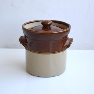 Puede incluir: Una vasija de cerámica cilíndrica con un acabado de dos tonos que presenta una sección superior esmaltada en marrón oscuro brillante y una sección inferior en beige claro mate. Incluye dos pequeñas asas laterales y una tapa a juego.