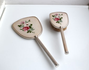 Puede incluir: Un conjunto vintage de espejo de mano y cepillo para el cabello plateado con diseños florales. El espejo y el cepillo tienen ambos un diseño floral con rosas rosas y blancas y hojas verdes.