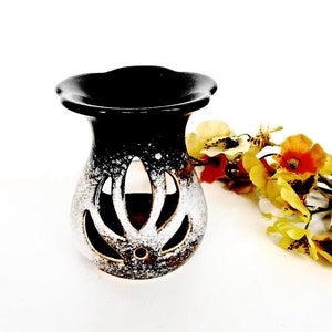 May include: A black and white ceramic oil burner with a decorative cutout design. The burner has a wide, flat top and a rounded base.