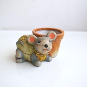 May include: A ceramic planter shaped like a gray mouse with orange ears and a yellow and brown vest. The planter has a brown clay pot on its back.
