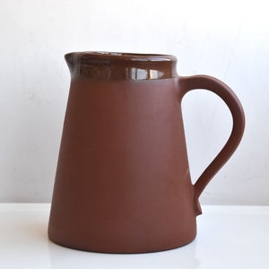 Peut inclure: Un pichet en terre cuite avec un bord émaillé brun et une anse. Le pichet est un design classique et serait parfait pour servir des boissons.