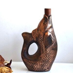 May include: A copper-colored ceramic bottle shaped like a fish with detailed scales. The bottle has a wide opening at the top and a handle formed by the fish's tail.