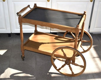 Puede incluir: Un carro de servicio de madera con dos niveles. El nivel superior tiene una superficie negra y el nivel inferior es de madera. El carro tiene cuatro ruedas, dos de las cuales son visibles en la imagen. El carro está en una habitación con gabinetes blancos en el fondo.