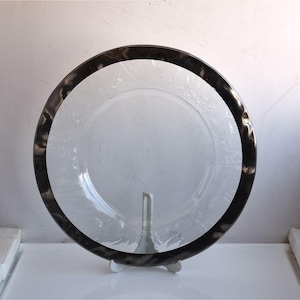 Puede incluir: Un plato de vidrio transparente con un borde oscuro y texturizado. El plato es redondo y se apoya en un soporte blanco. El borde presenta un patrón arremolinado, agregando un elemento decorativo. El plato es probablemente para servir alimentos.