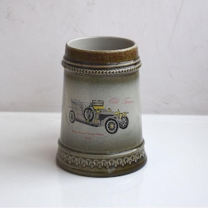 May include: A ceramic beer stein with a vintage car illustration. The stein has a light green and brown color scheme, with a decorative base and a braided design around the top. The car graphic is labeled "Old Timer" and "Rolls-Royce Silver Ghost 1907".