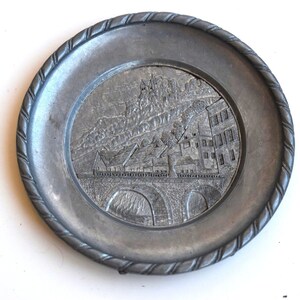 May include: A silver metal plate with a raised design of a bridge and buildings. The plate has a rope-like border.