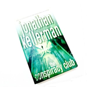 Può includere: Una copertina di libro verde e bianca con il titolo "The Conspiracy Club" di Jonathan Kellerman.