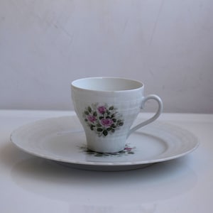 Peut inclure: Une tasse à thé et une soucoupe en porcelaine blanche avec un motif floral représentant des roses roses et des feuilles vertes.
